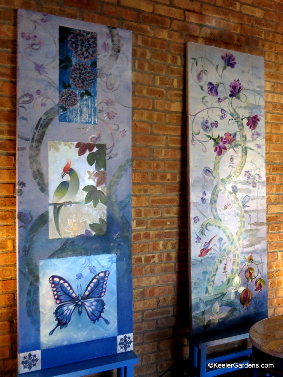 Two tall canvas paintings by Meg Fine Art, sit side by side at an art show. Heavy blue tones cover both with as the one on the left depicts a vining flower with accents of a red cardinal and the one on the right features a beautiful blue butterfly.