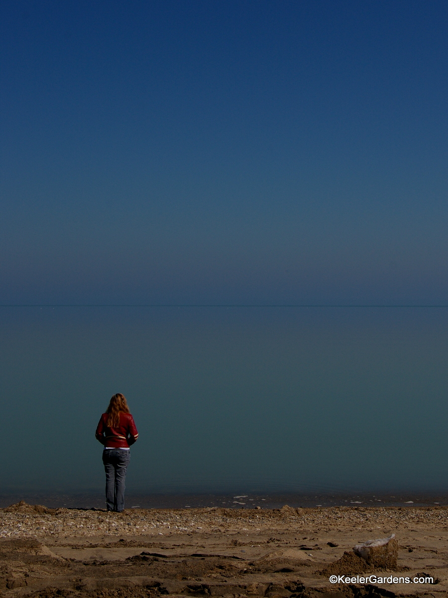 A lone visitor to a somewhat rocky beach in Lake Bluff, IL stands near the water's edge on peers out to an apparently infinite expanse that is Lake Michigan that at some point meets the sky in a thin blue line.