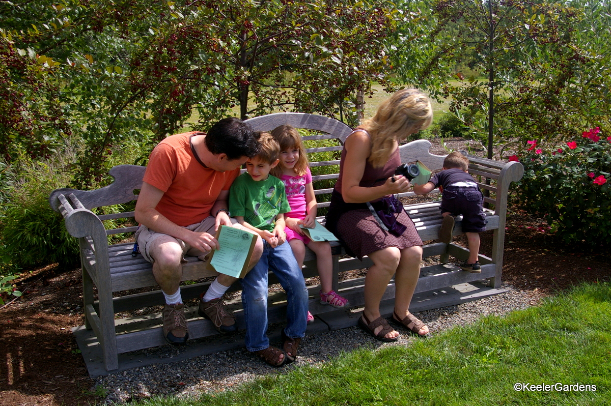 Two adults sit on a bench in a display garden with three children. The man works with a boy and girl of 5 and 6 years old. The women with her camera keeps an eye on the 3 year old who is more interested in climbing the bench.