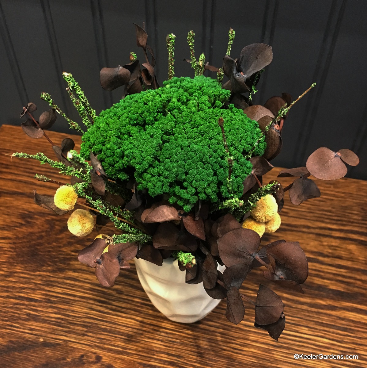A sweet cluster of dried botanicals including green yarrow, chocolate-colored eucalyptus, yellow flower buttons, and green-flocked birch branches, all in a miniature off-white rippled vase.