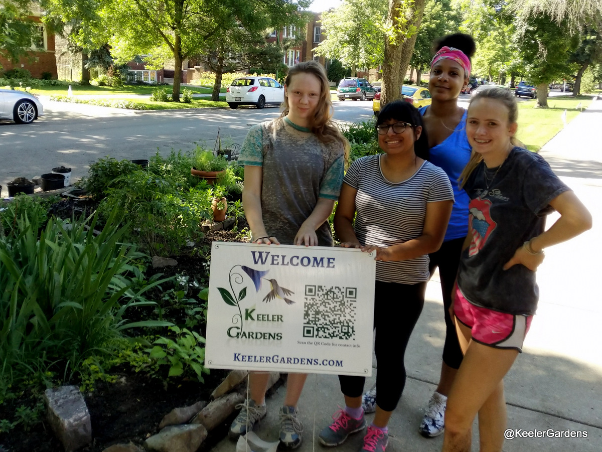 Four high school interns from across Chicago pose for a picture, standing in the right foreground in a group on a sidewalk with a sign that reads “Welcome, Keeler Gardens.” In the background, mostly to the left, is the Keeler Gardens educational pollinator habitat, freshly planted, with a variety of Illinois native pollinator plants.