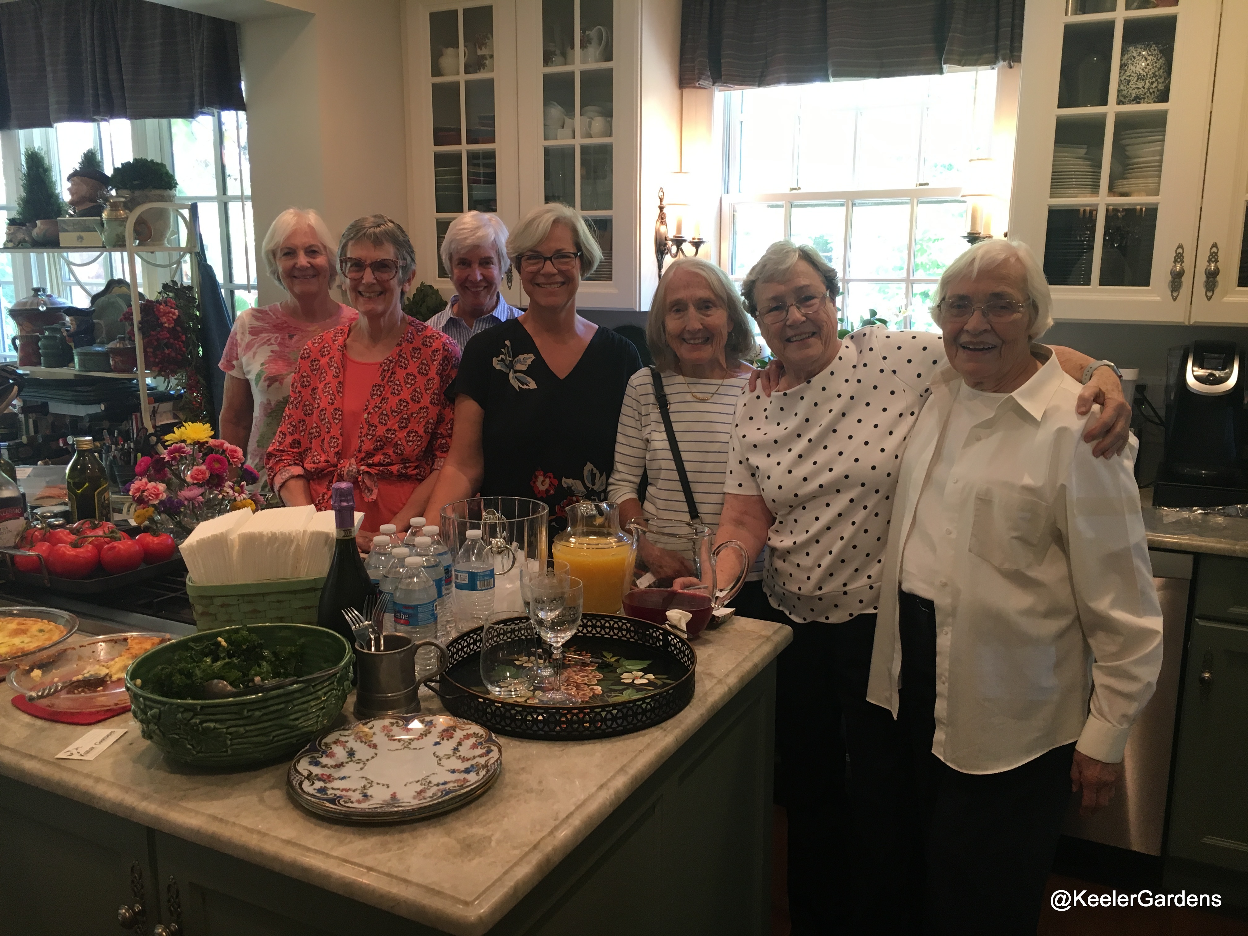 A group of ladies poses for a picture in a well appointed kitchen. On the island there is a bountiful display of food and beverage.